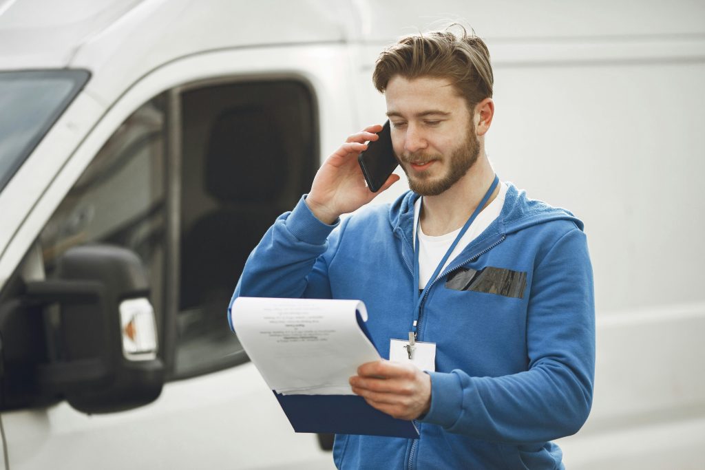 A delivery man in a blue hoodie makes a phone call while holding a clipboard, standing by a white van.